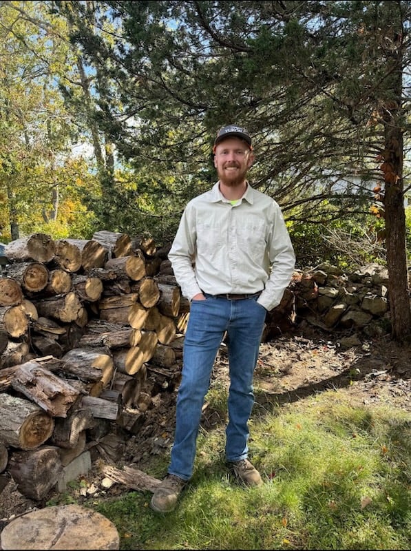 A picture of our owner, Weylon Miller, standing next to a neatly stacked cord of firewood.